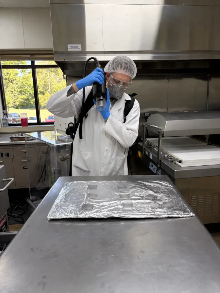 Technician in PPE using a SteraMist SteraPak handheld applicator to apply ionized hydrogen peroxide mist to stainless steel test coupons in a food lab